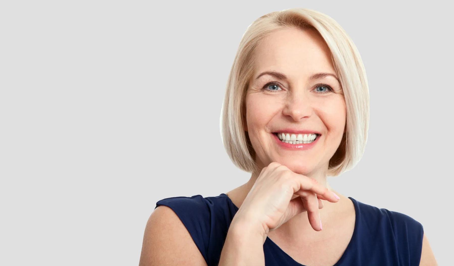 Smiling middle-aged woman with short blonde hair resting her chin on her hand against a gray background.