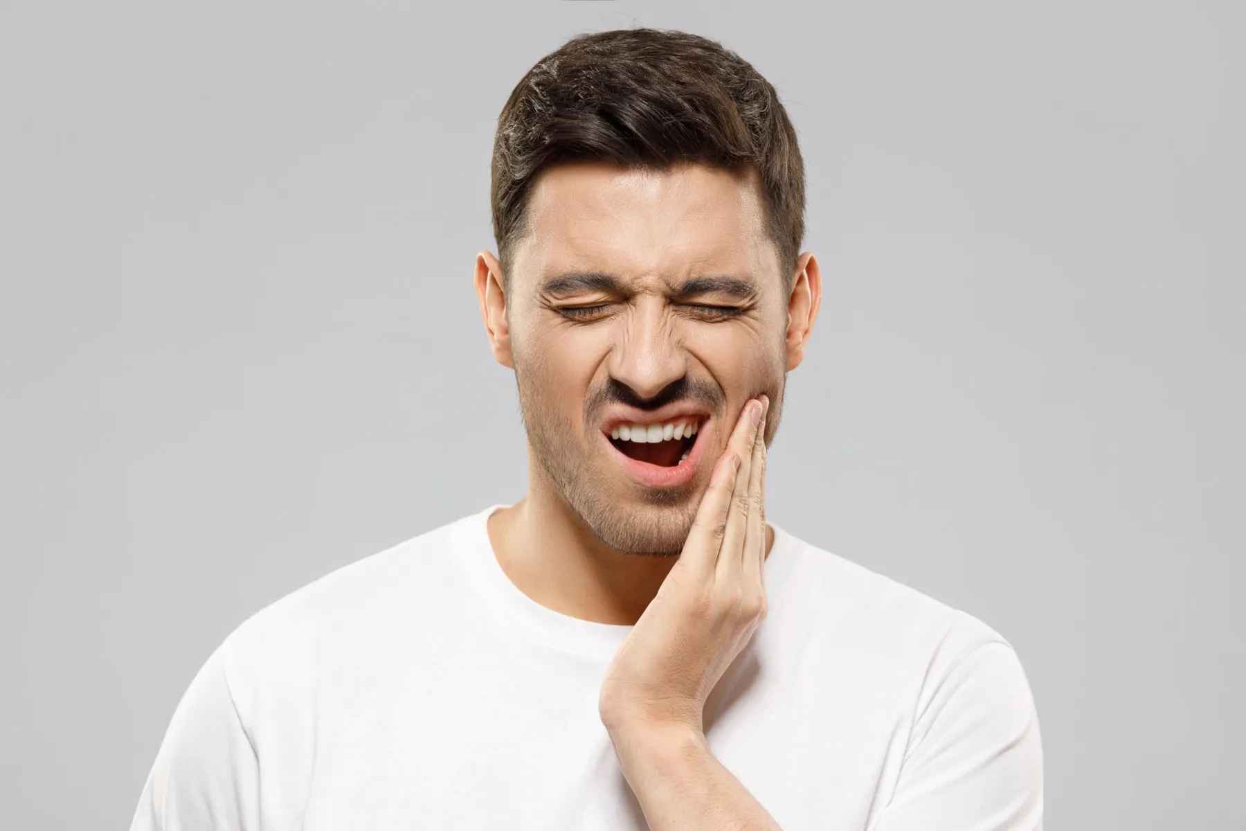 Man in white shirt holding his cheek with a pained expression, indicating toothache or jaw pain.