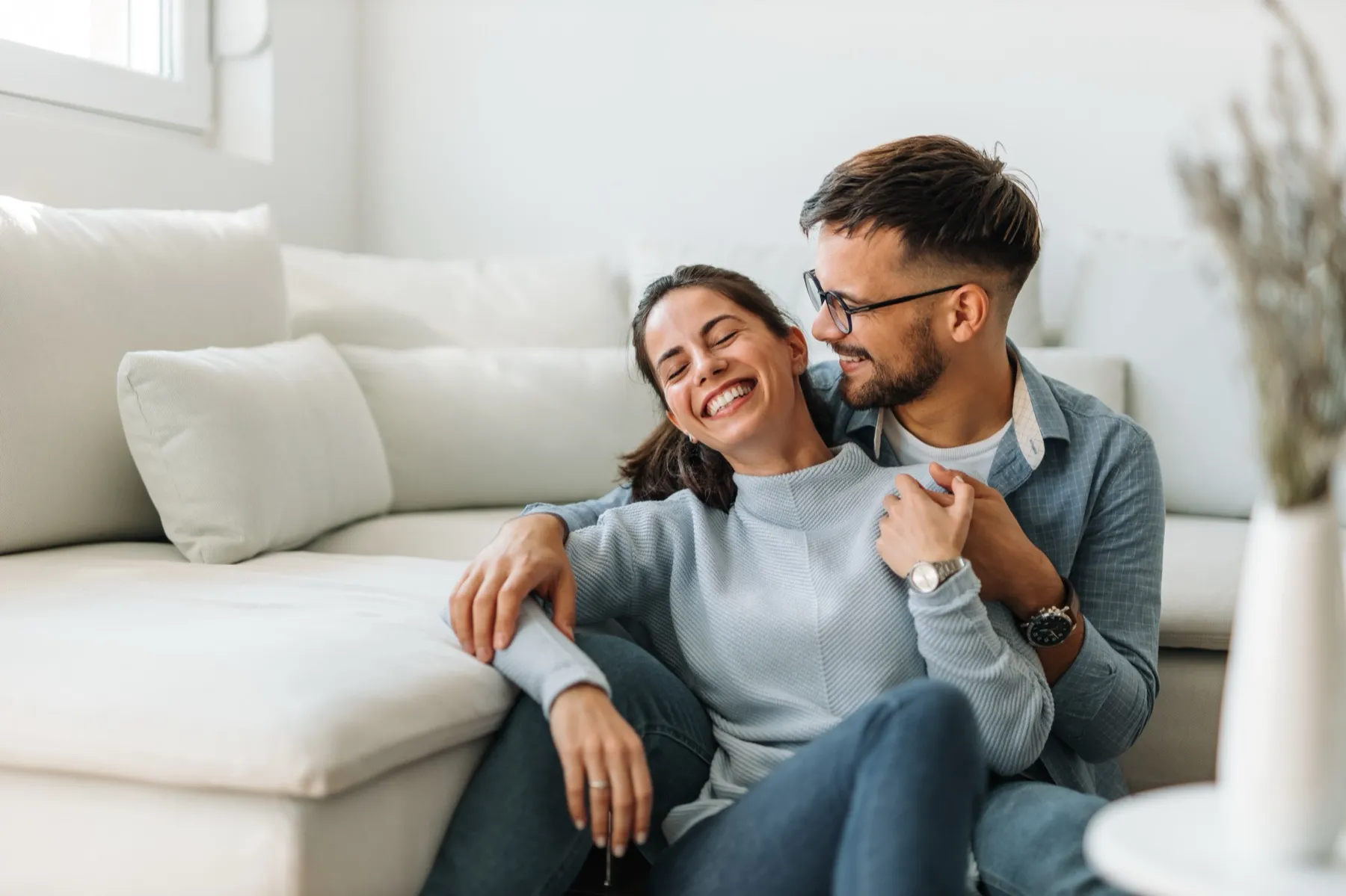 Smiling couple sitting on the floor in a bright living room, embracing near a white couch.