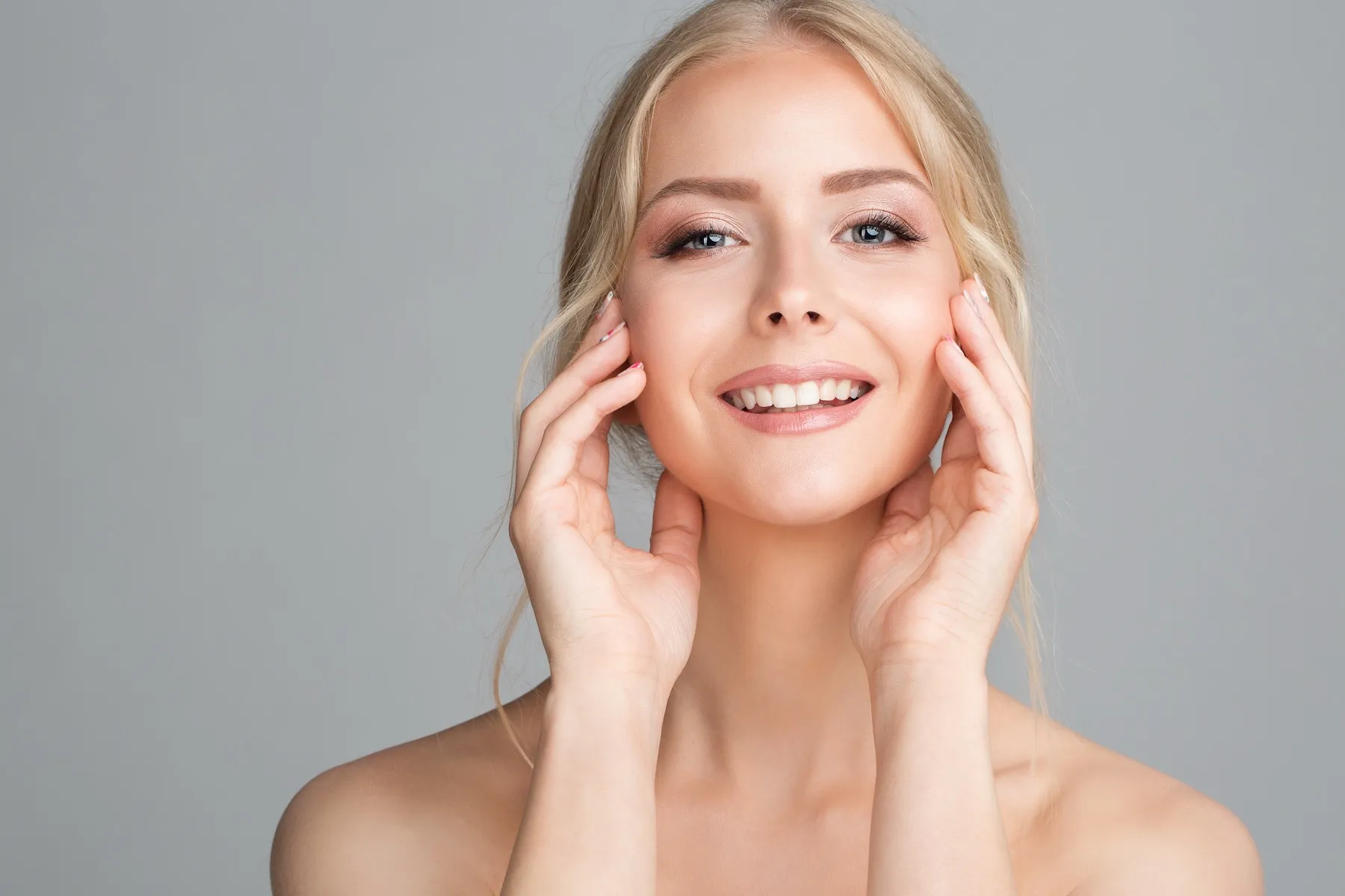 Smiling blonde woman with clear skin gently touching her face against a light grey background.