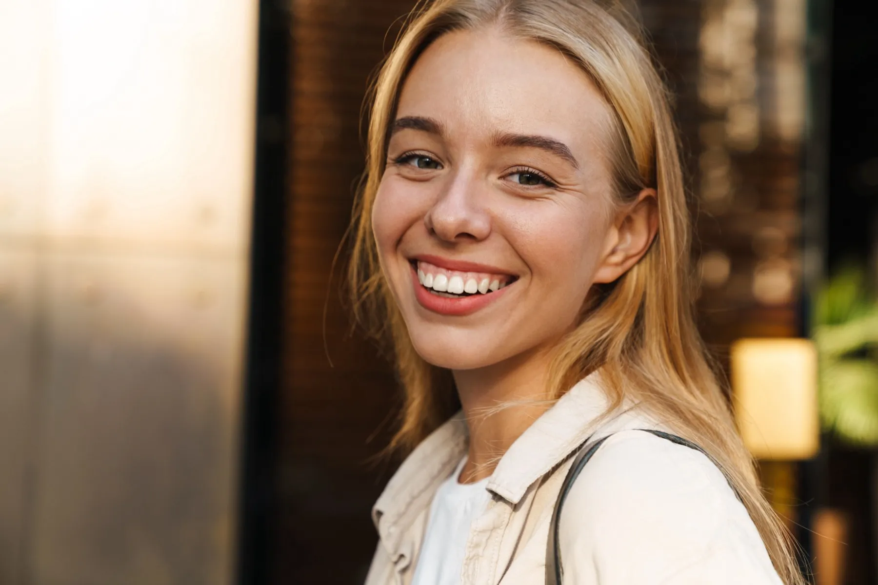 Smiling young woman with blonde hair wearing a light-colored jacket, standing indoors with softly blurred background.