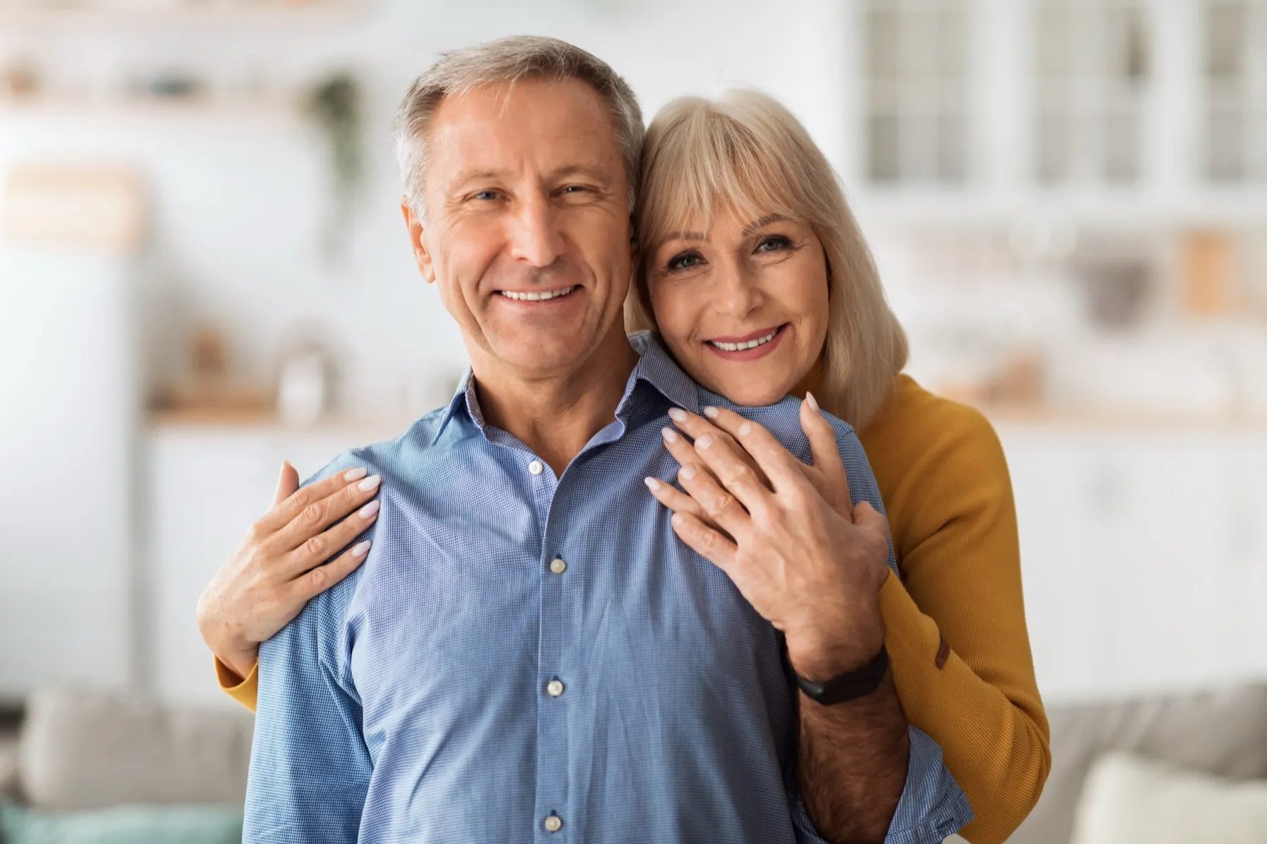Smiling senior man in blue shirt being embraced from behind by a cheerful senior woman in a mustard sweater in a bright home setting.