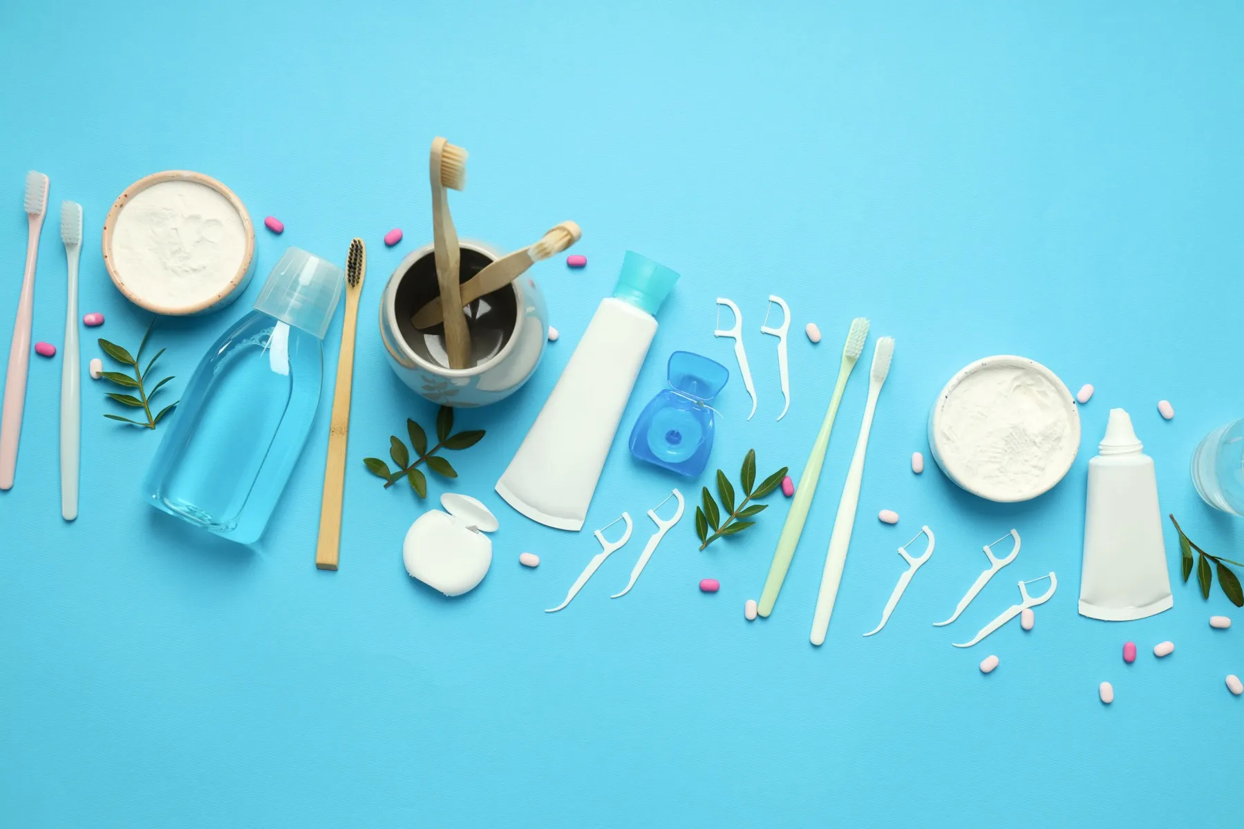 Various dental care items including toothbrushes, dental floss, toothpaste, toothpowder, mouthwash, and floss picks arranged on a blue background with green leaves and pink and white tablets.