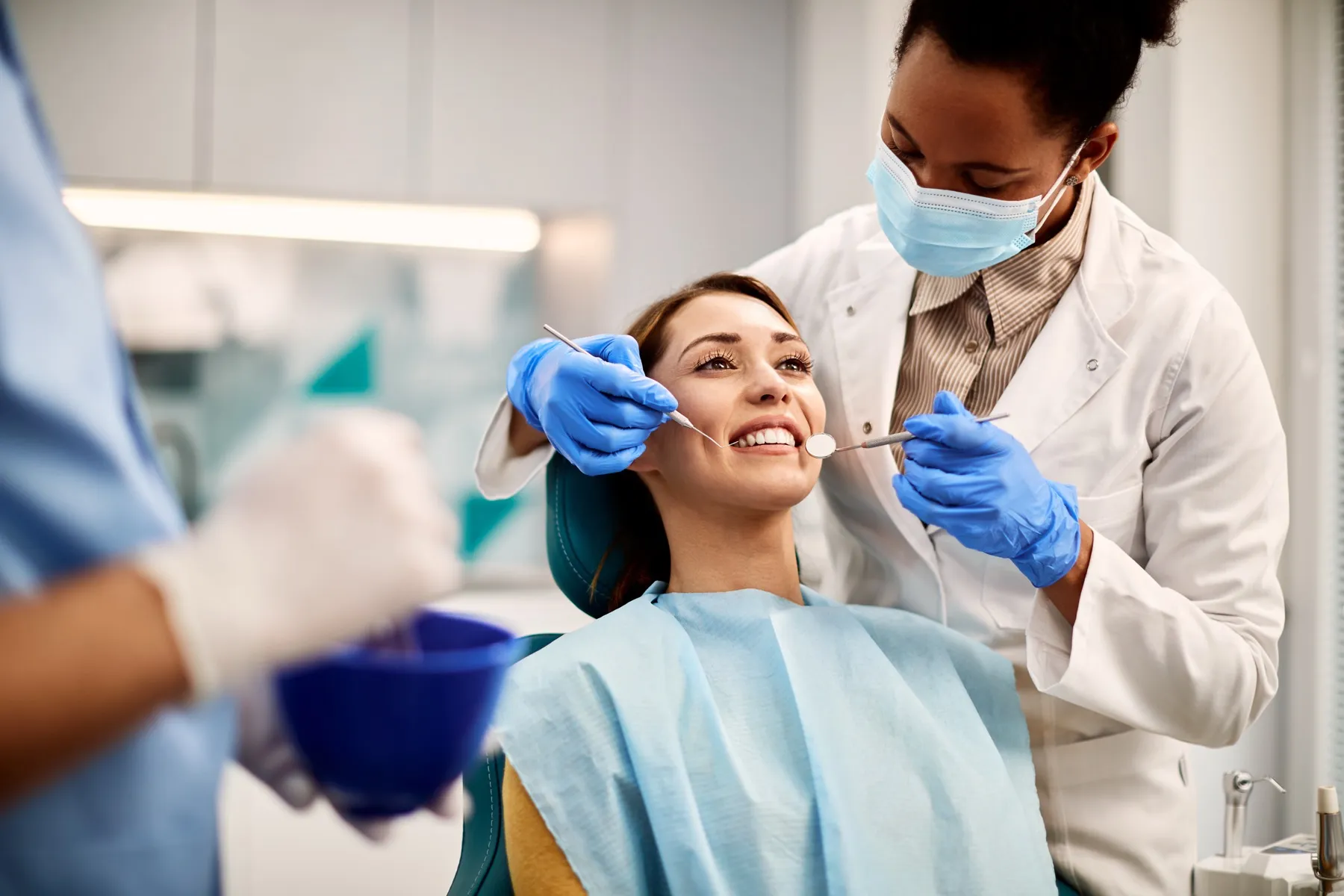 Dentist wearing blue gloves and a mask examining a smiling female patient's teeth with dental tools in a clinic.
