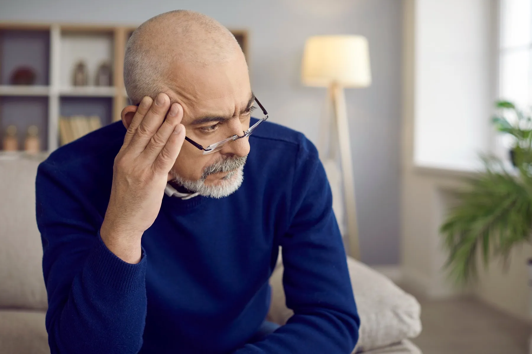 Pensive older man with glasses resting his head on his hand while sitting in a living room.