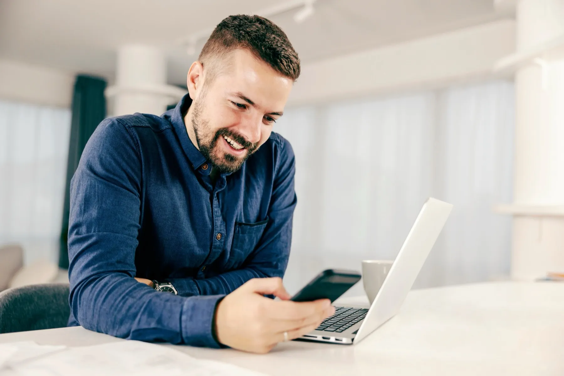 Smiling man in blue shirt sitting at a table, looking at his phone with a laptop and coffee cup nearby.