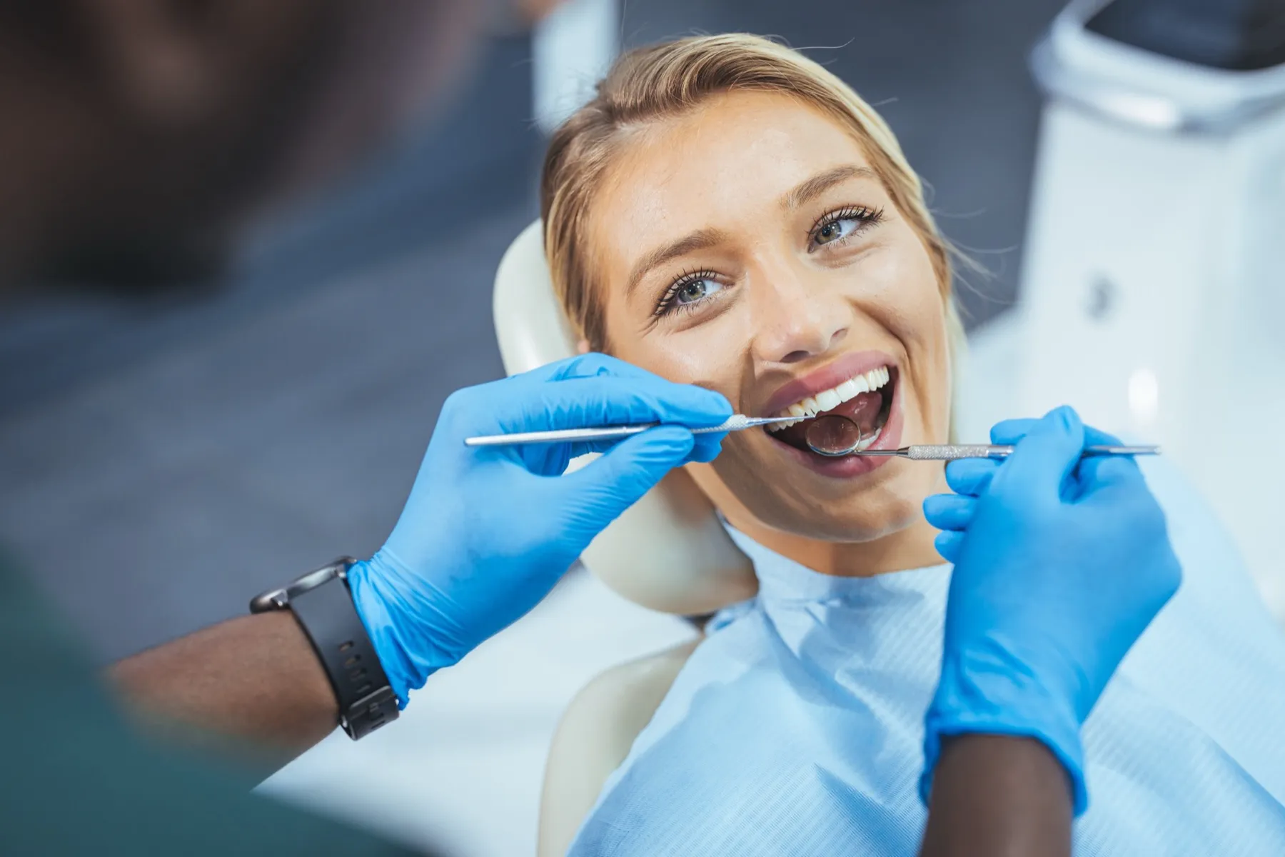 Dentist wearing blue gloves examining a smiling woman's teeth with dental tools.