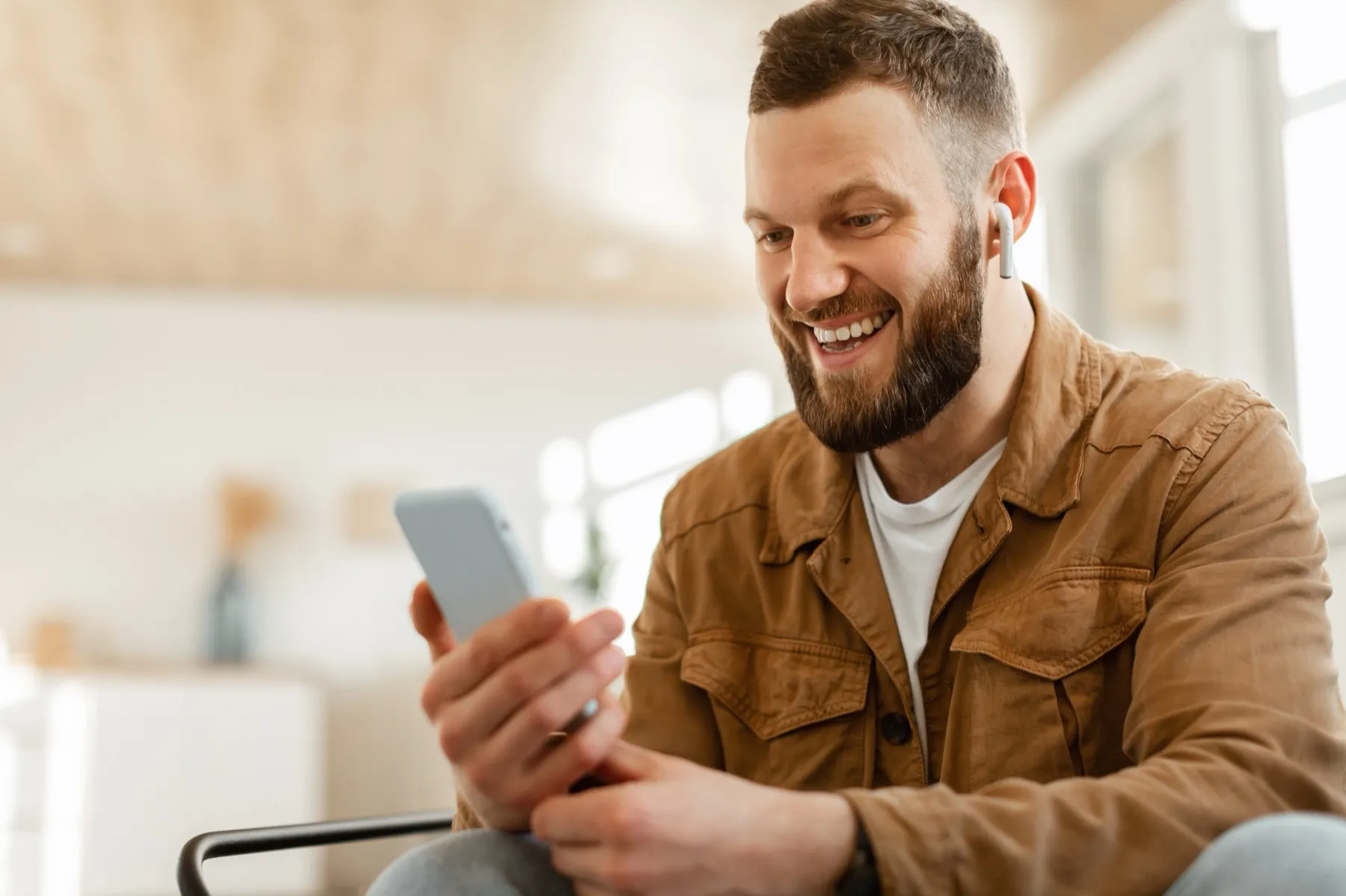 Smiling man with a beard wearing a brown jacket and white earbuds holding a smartphone.