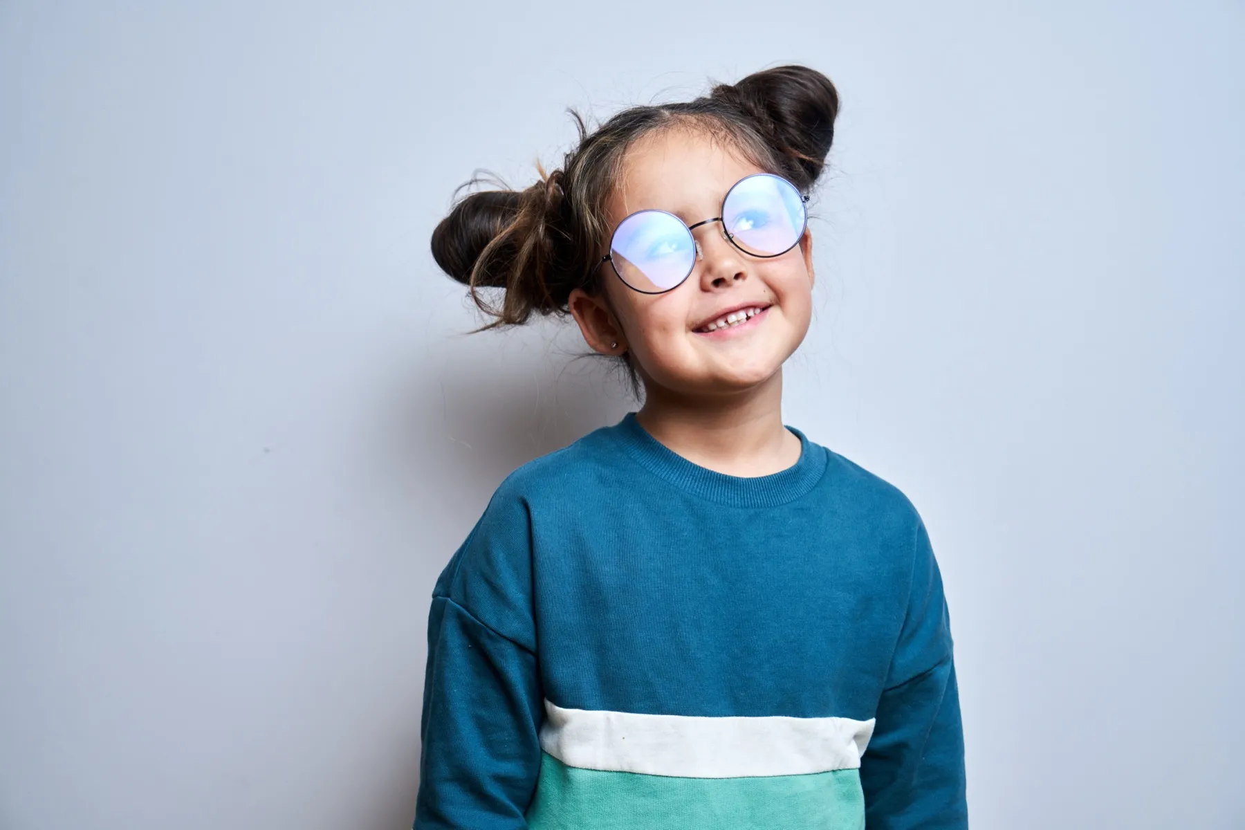 Smiling young girl with round glasses and two buns, wearing a blue and green color-block sweater against a plain light background.