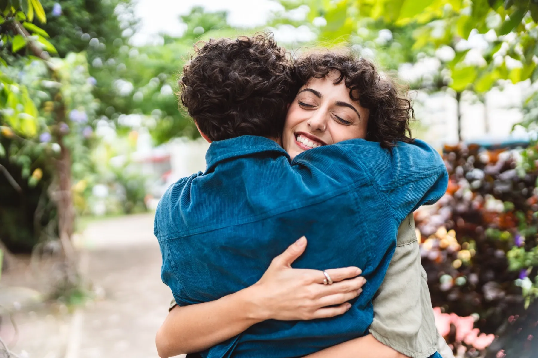 Two people with curly hair warmly hugging outdoors amid greenery.