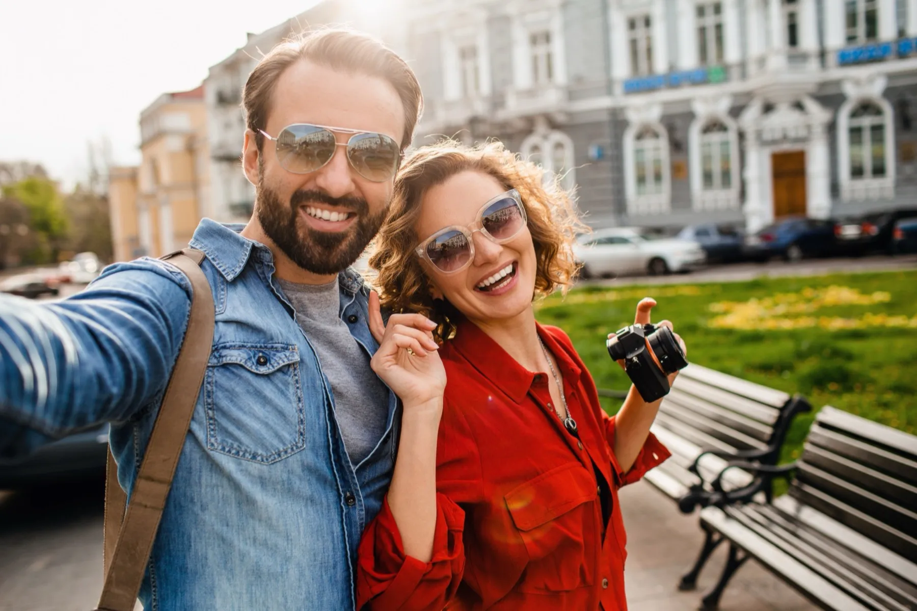 Smiling man and woman wearing sunglasses taking a selfie outdoors with a camera, benches, and building in the background.