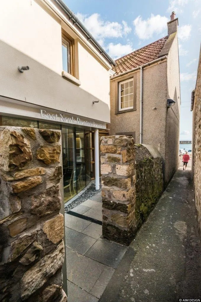 Narrow alleyway between stone walls leading to an entrance with glass doors of East Neuk Dental Practice building under a partly cloudy sky.