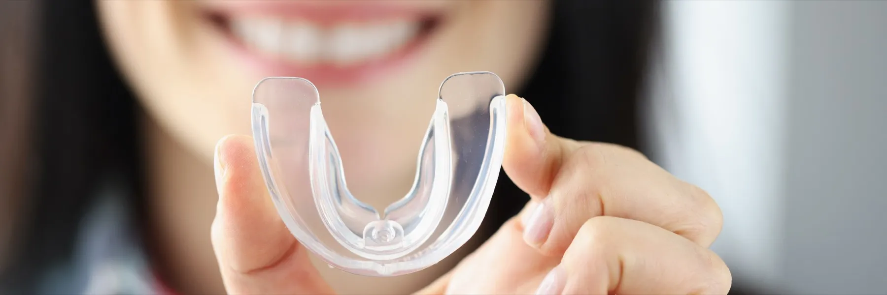 Person holding a transparent mouthguard with a blurred smiling face in the background.