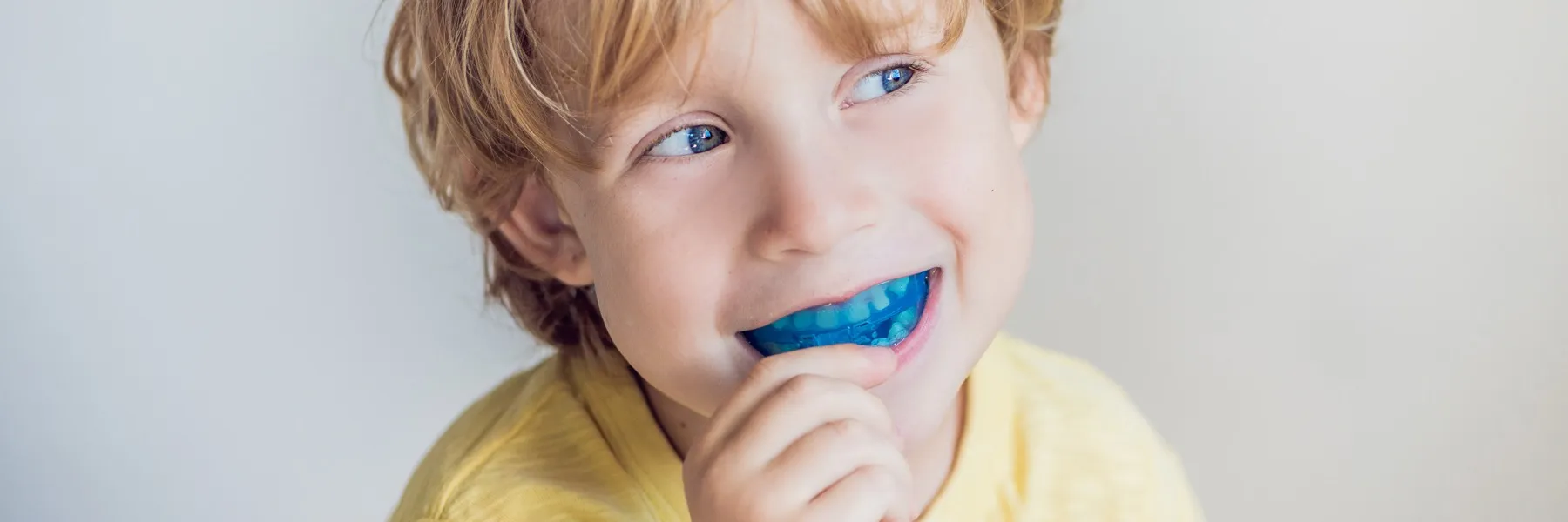 Young boy with blue eyes holding and smiling with a blue mouthguard in his mouth.