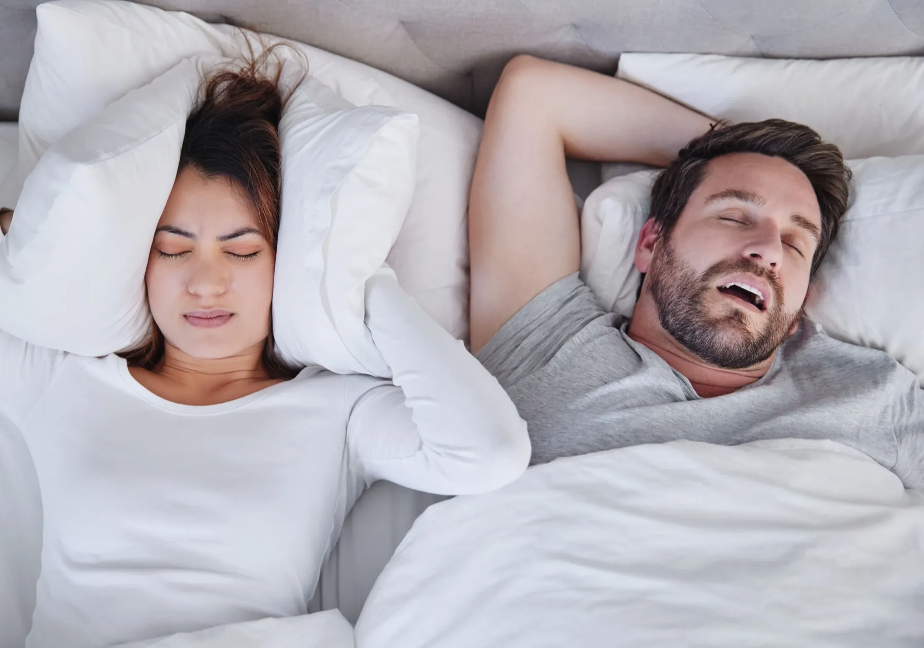 Woman covering her ears with pillows annoyed by man snoring beside her in bed.