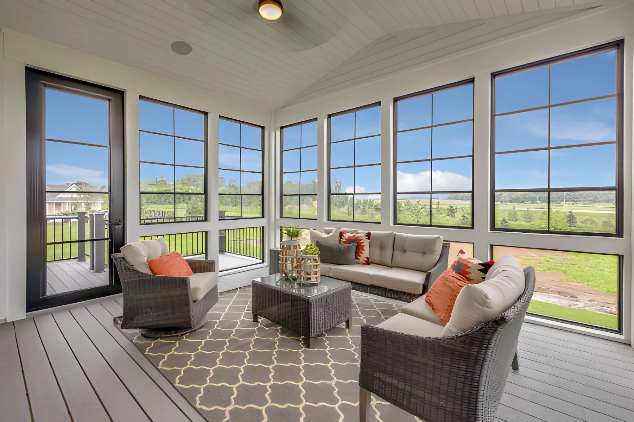 Sunroom with large grid windows overlooking green fields, furnished with wicker chairs, sofa, and a central table on a patterned rug.