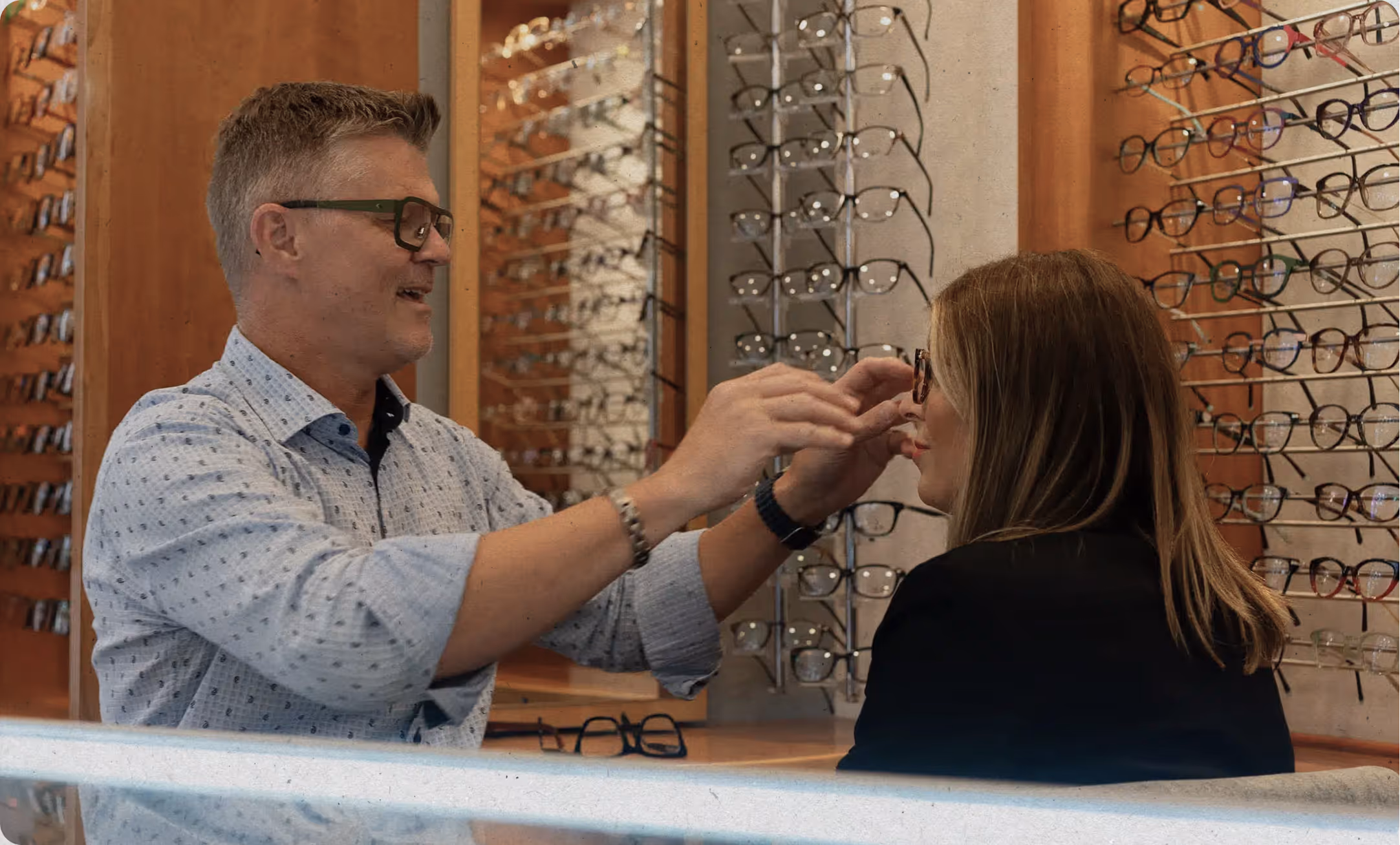 Optician fitting eyeglasses on a woman inside an eyewear store with multiple glasses displayed on wall racks.