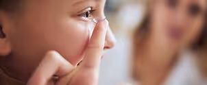 Close-up of a person inserting a contact lens into their eye with another person blurred in the background.