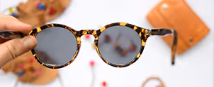 Close-up of a hand holding round tortoiseshell sunglasses with blurred accessories in the background.