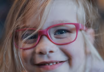 Close-up of a smiling child with blonde hair wearing pink eyeglasses.