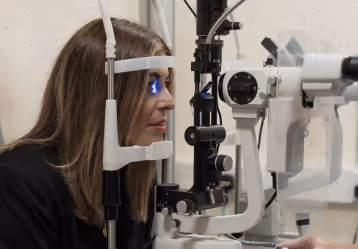 Woman undergoing an eye examination using a slit lamp device in a clinical setting.