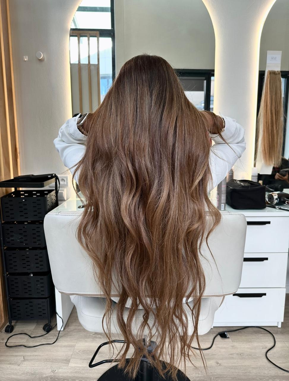 Person with long, wavy light brown hair sitting in a salon chair facing a mirror.