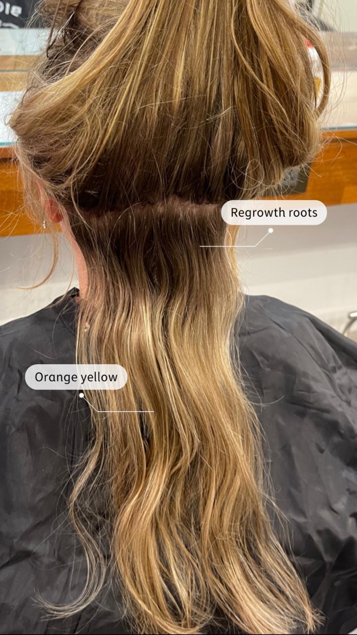 Back view of a woman's hair showing darker regrowth roots beneath lighter orange-yellow dyed hair.