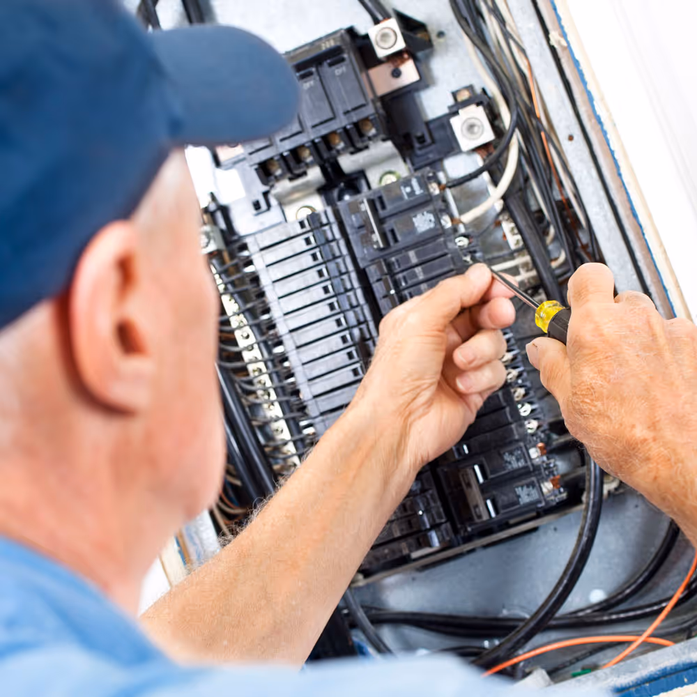 Das Bild zeigt eine Person in Arbeitskleidung, die an einem elektrischen Schaltschrank arbeitet und dabei Kabel oder Bauteile überprüft
