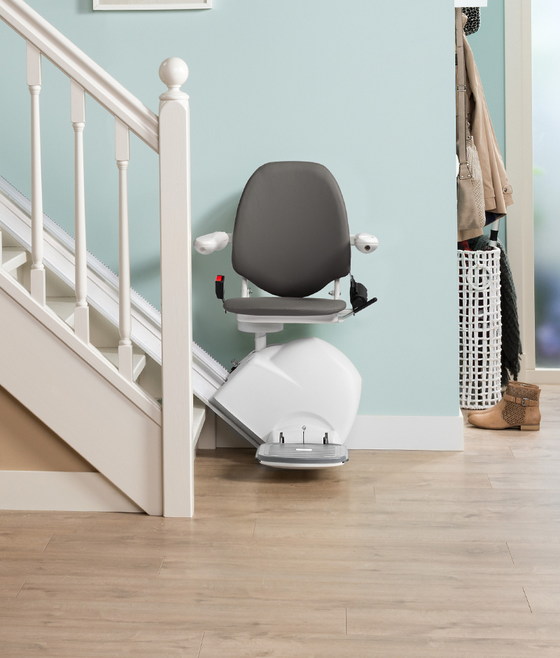A modern, minimalist bathroom features a white staircase with a black seat and footrest, a white toilet with a black seat and footrest, a white handrail, a wooden floor, a white basket, and a coat rack, all set against a light blue wall.