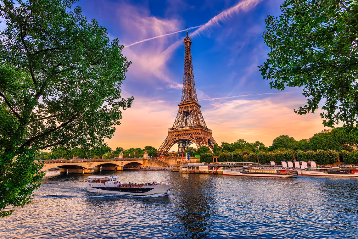 Eiffel Tower in Paris at sunset with river Seine and boats in the foreground framed by leafy green trees.