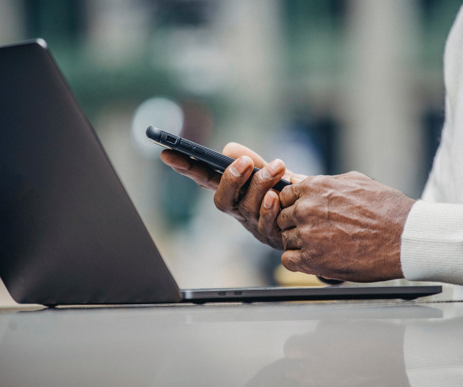 Close-up of a person's hands holding a smartphone near an open laptop on a table.