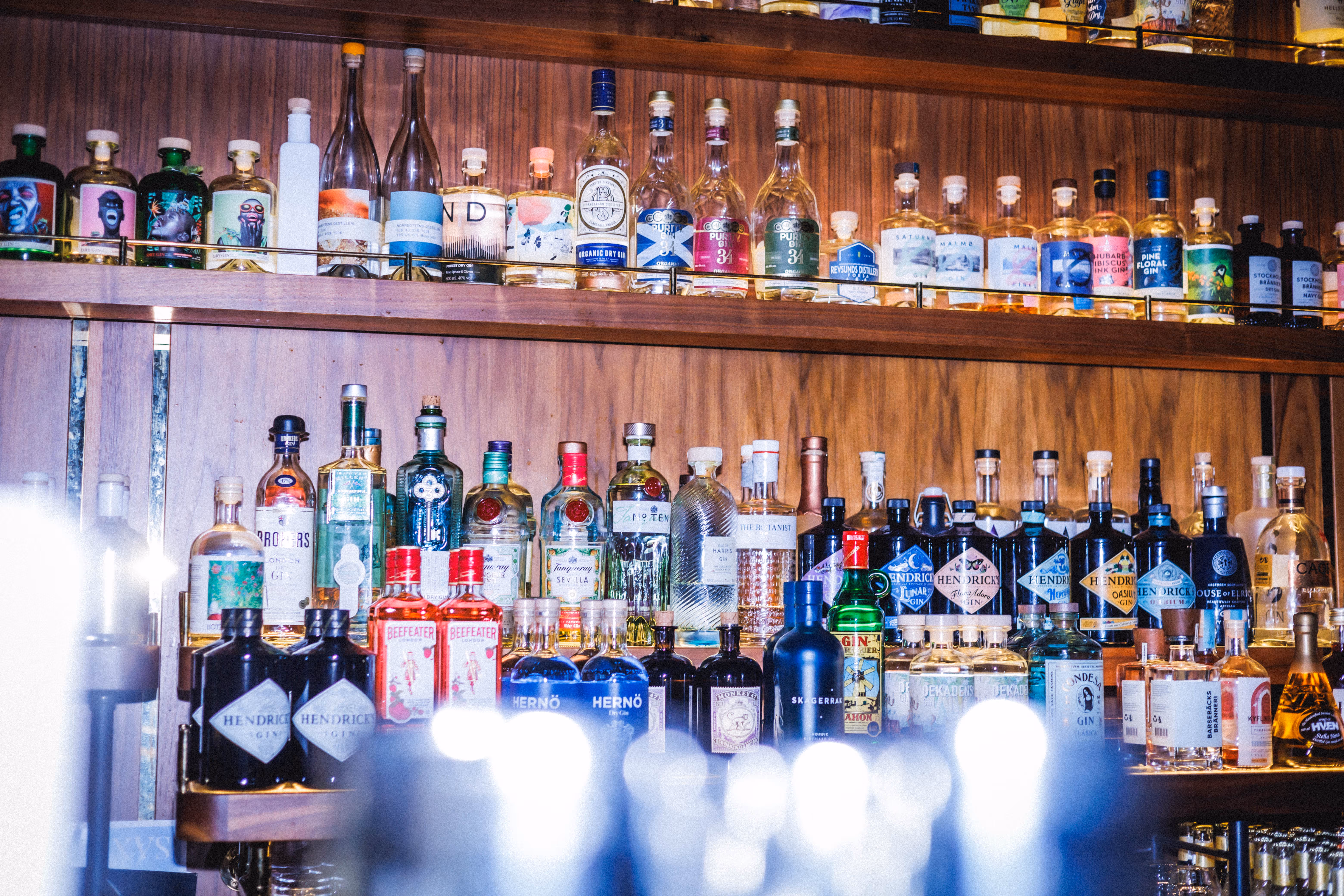 Hylla fylld med över 300 ginflaskor i olika former och färger framför en träpanelvägg.