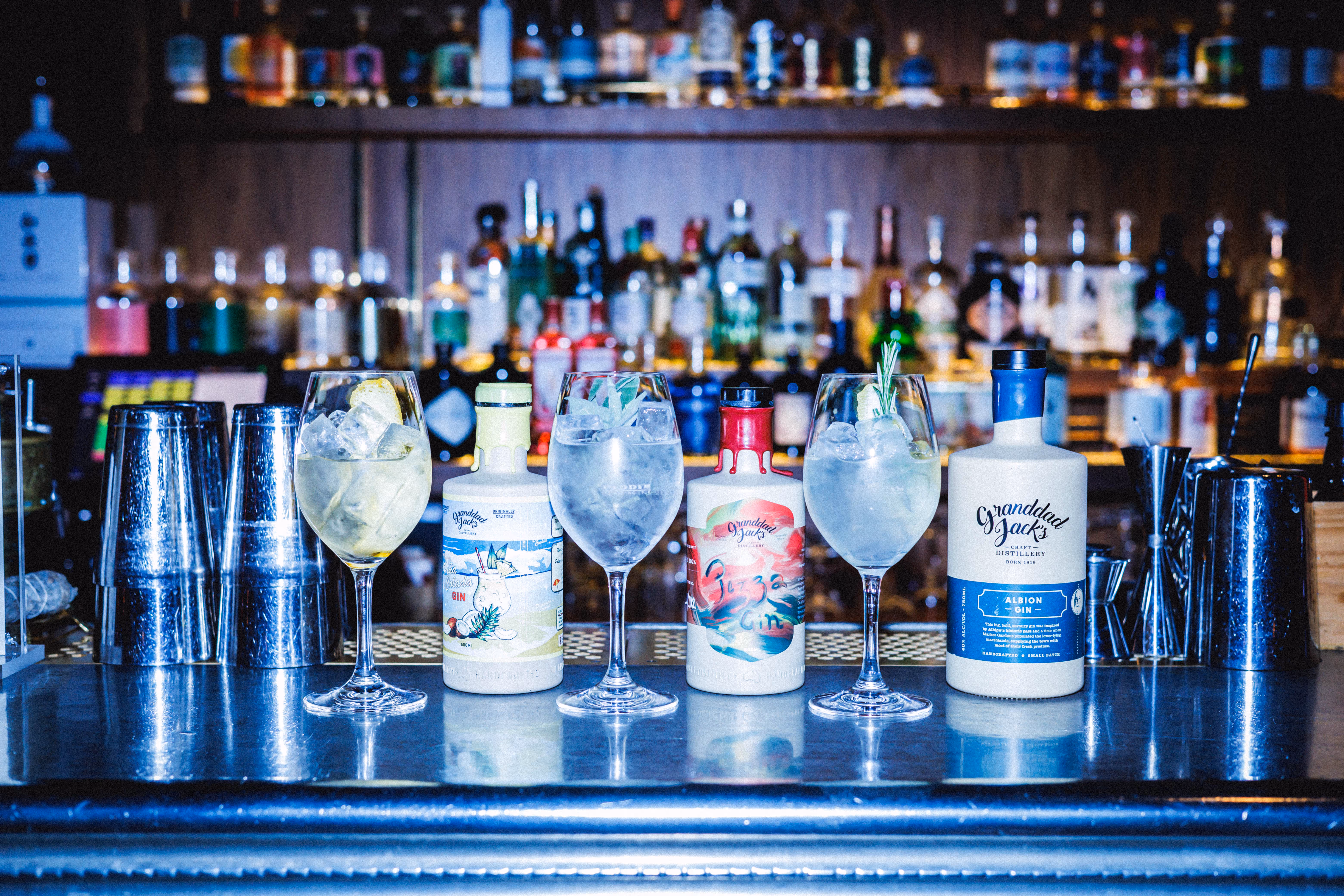 Tre flaskor Granddad Jack's gin med tre glas fyllda med is och garnityr på en bar med flaskor i bakgrunden.