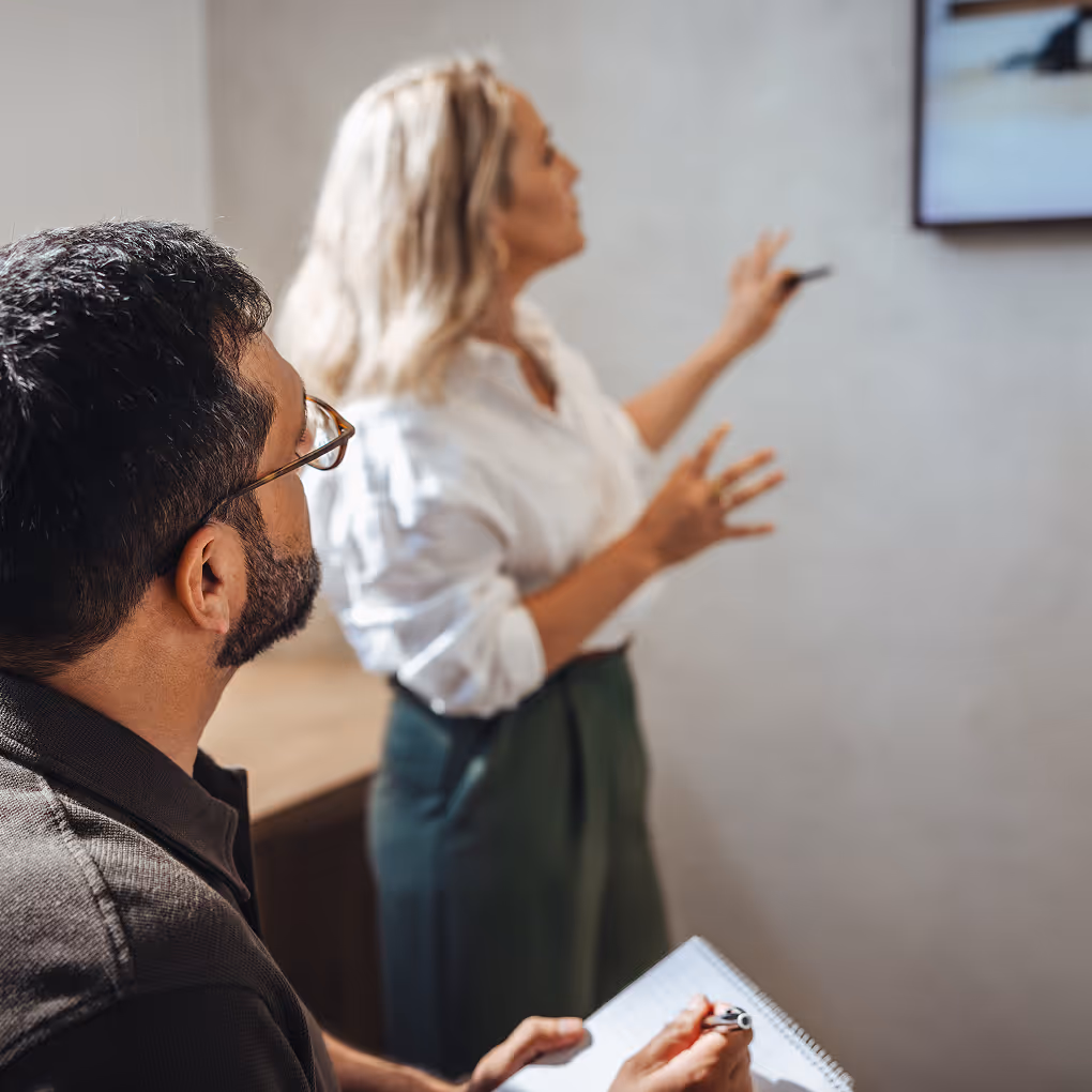 Eine Frau präsentiert vor einem Bildschirm, während ein Mann mit Brille Notizen macht.