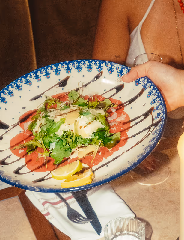 Servieren eines Carpaccio-Tellers im Italian Shot Glockenbach, umgeben von Gästen und Kerzenschein.