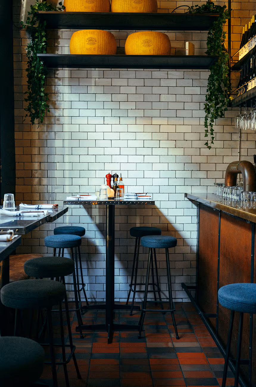 Hochtische und Barhocker im Italian Shot Maxvorstadt vor weiß gefliester Wand und großen Parmesanlaiben.