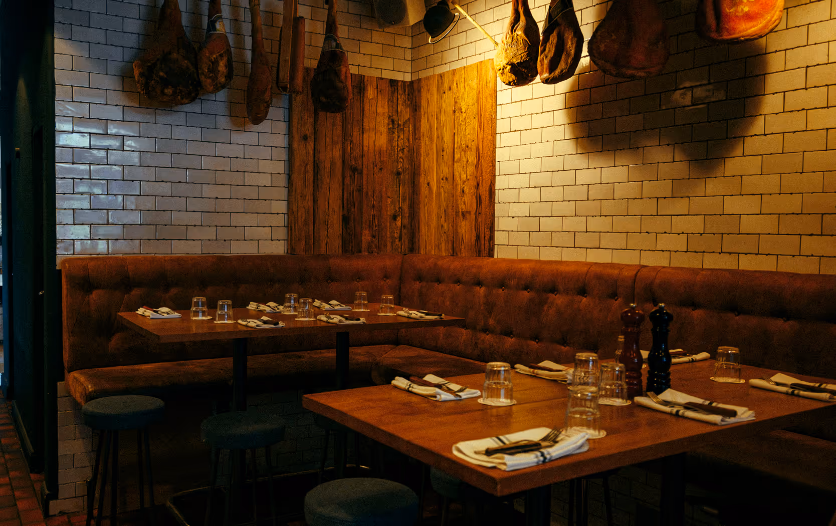 Eckbereich im Italian Shot Maxvorstadt mit Lederbank, gedeckten Holztischen und dekorativ aufgehängten Schinken.