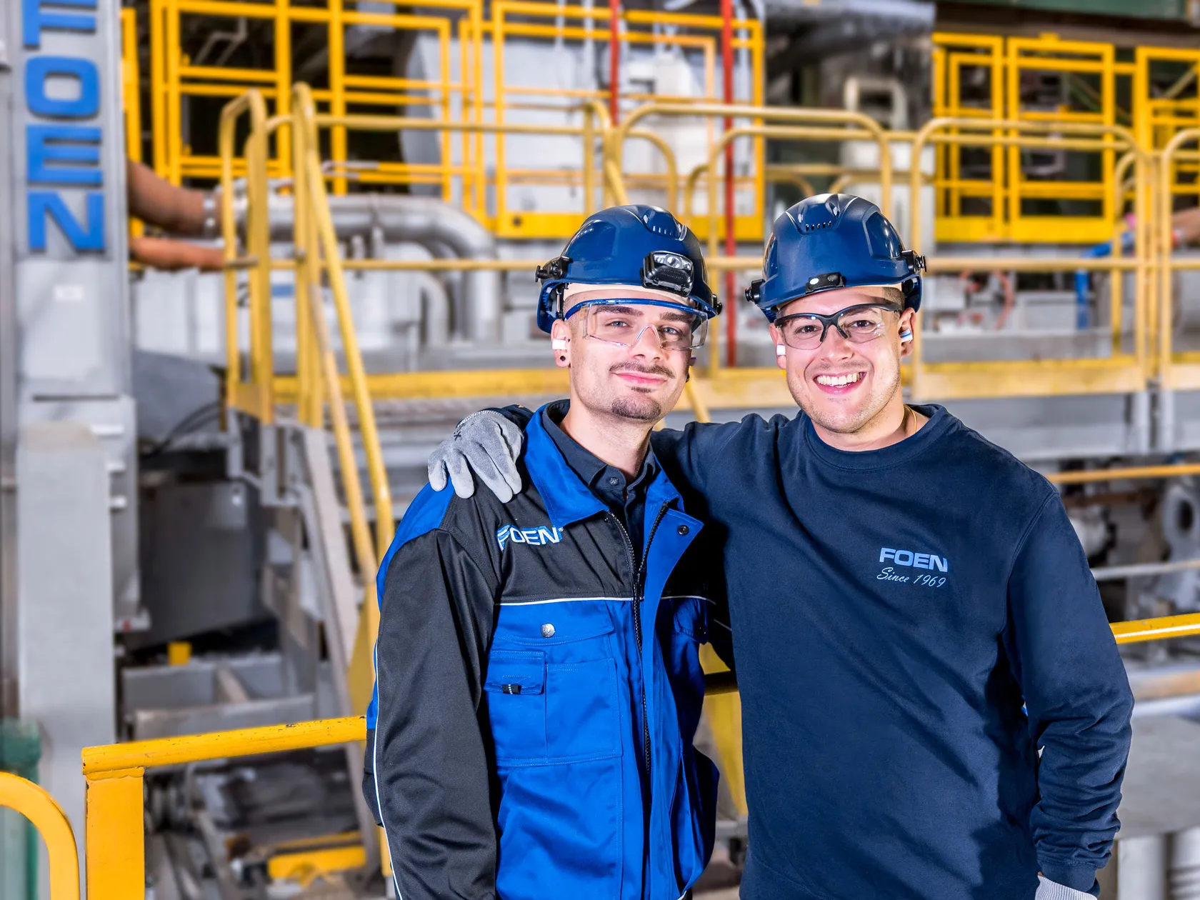 Zwei lächelnde Industriearbeiter mit Schutzhelmen und Brillen posieren in einer Fabrikumgebung mit gelben Geländern.