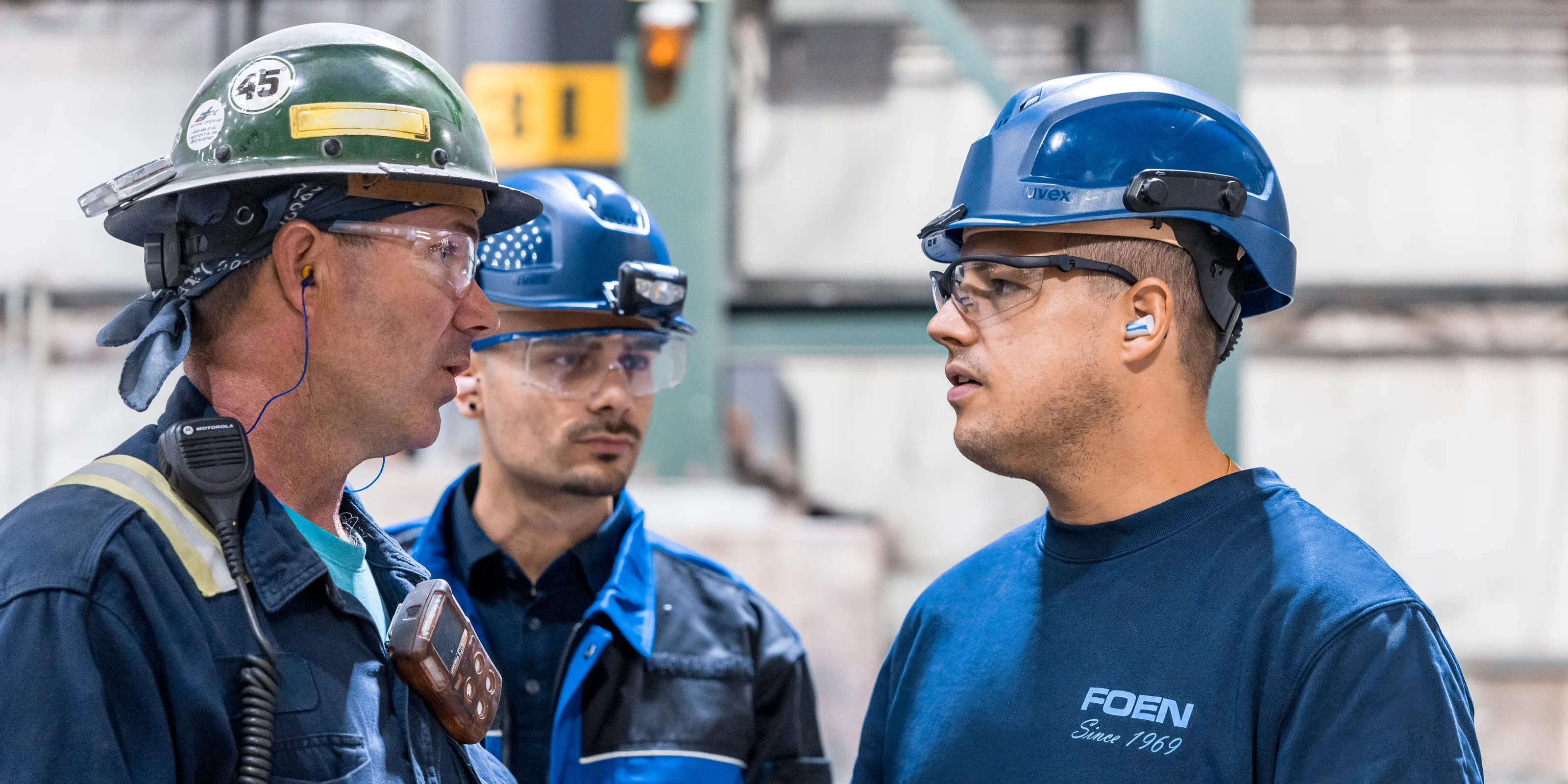 Drei Industriearbeiter mit Helmen und Schutzbrillen im Gespräch in einer Fabrikumgebung.