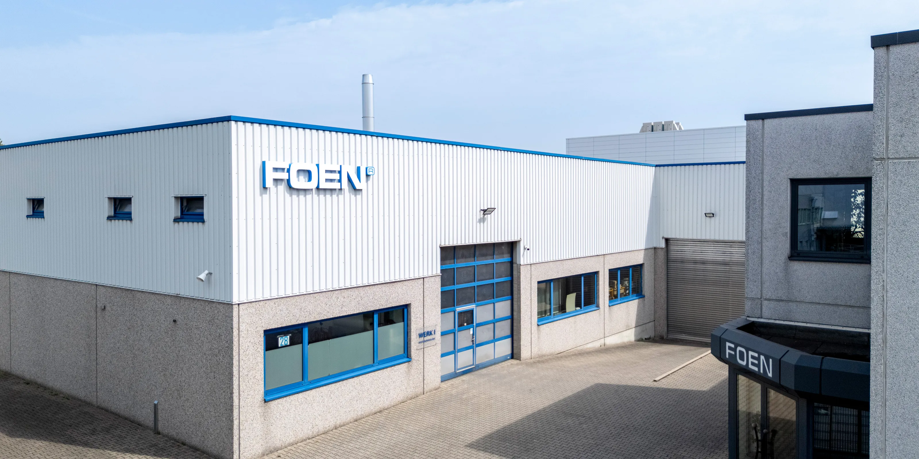Industrial factory buildings with FOEN logo on white and grey façade under blue sky.