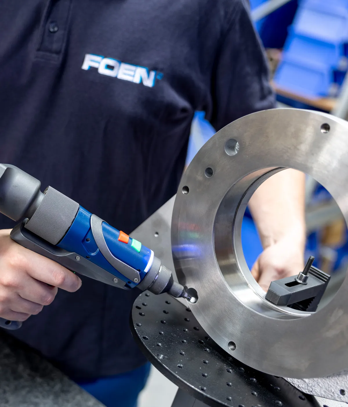 Craftsman in FOEN polo shirt uses blue electric screwdriver on large round metal part.