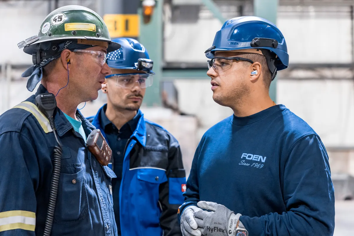 Three factory workers wearing safety helmets and goggles talking in an industrial environment.