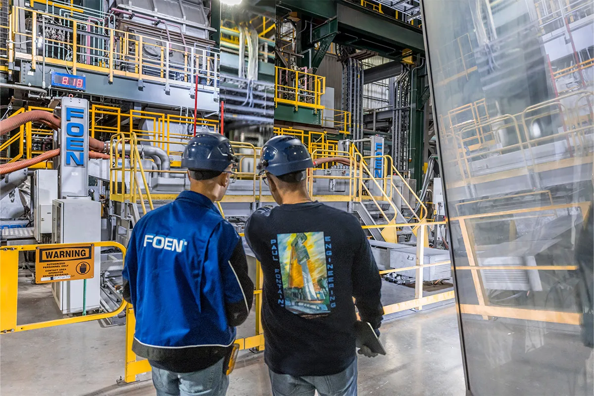 Two workers wearing blue helmets and hair dryers are standing in an industrial plant and discussing something.