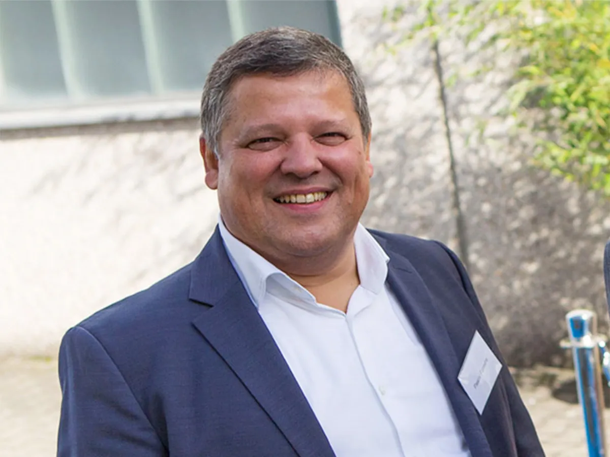 Smiling man in dark blue suit and white shirt, outside against a blurred background.