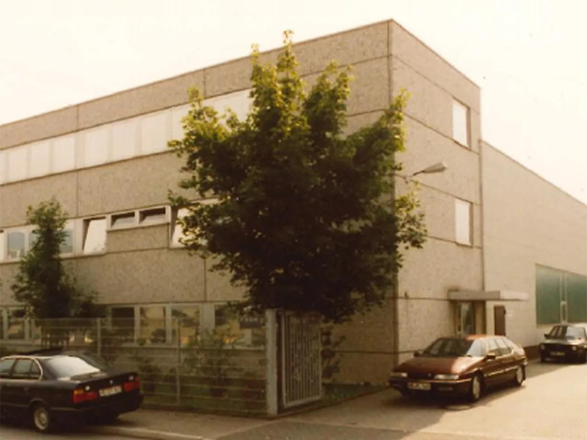 Older, two to three-story office building with grey concrete façade, trees in front of it and three parked cars.