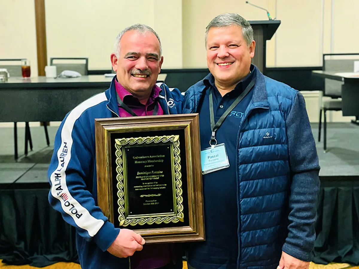Two men smile and hold a framed award, one wearing a Polo Ralph Lauren suit, the other a blue quilted coat.