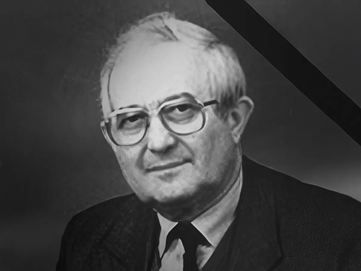 Black and white portrait of an elderly man wearing glasses and a suit against a dark background.