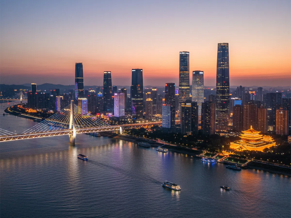 Städtische Skyline bei Sonnenuntergang mit beleuchteter Brücke über einen breiten Fluss und mehreren Booten auf dem Wasser.