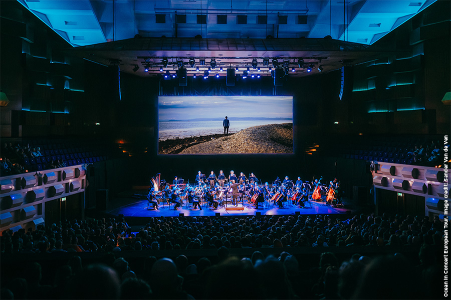 Ocean in Concert on a large screen with an orchestra playing in a concert hall while an audience looks on