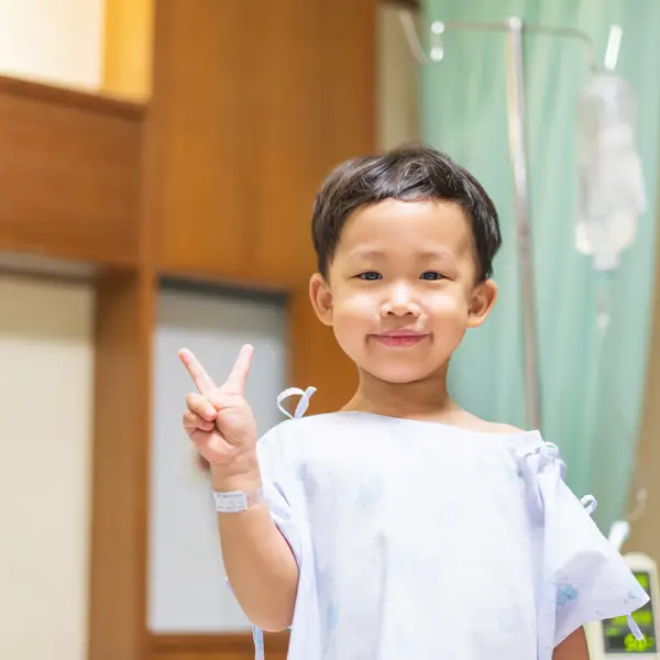 Smiling young child in a hospital gown showing a peace sign with a medical IV drip in the background.